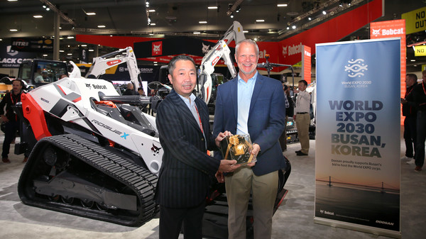 박정원 두산그룹 회장이 미국 라스베이거스에서 열린 ‘콘엑스포 2023’의 두산밥캣 부스에서 무인 전기 콘셉트 로더 ‘로그 X(Rogue X)’ 와 부산엑스포 유치를 기원하는 배너 앞에서 마이크 볼웨버 북미 지역장에게 두산일두(斗山一斗)를 전달하고 있다. 사진=두산 제공